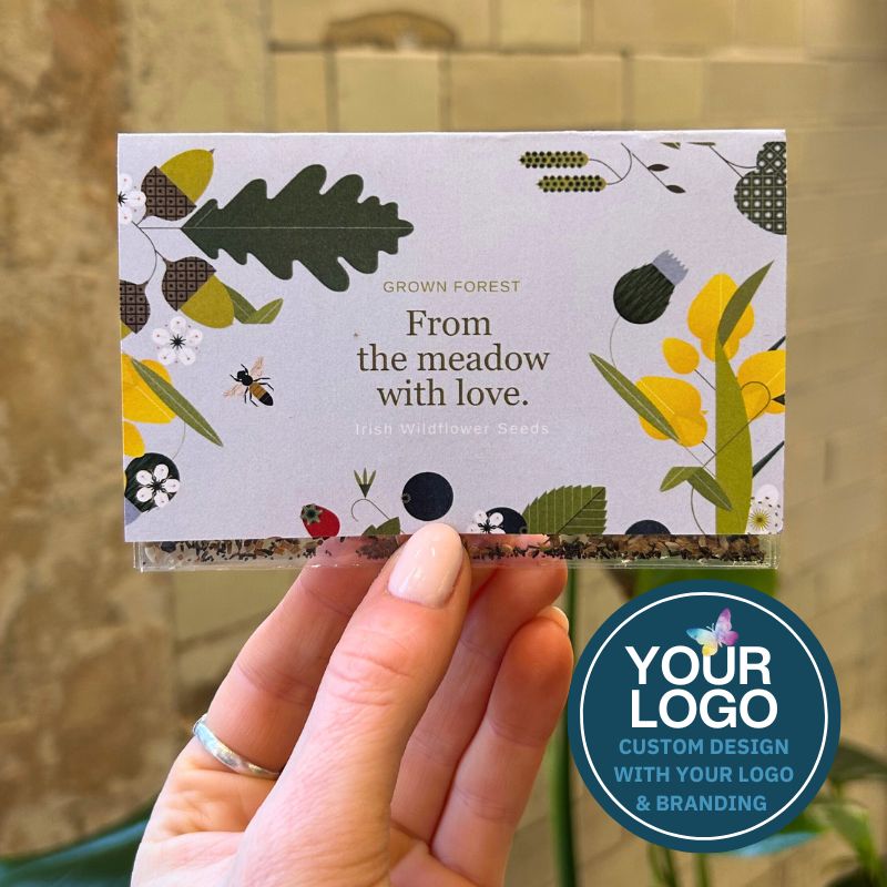 A corporate seed favour held in a hand, showing the front of the card with an illustrated forest scene, company logo and the text 'From meadow with love'. Overlaid text reads 'Your logo - custom design with your logo and branding'
