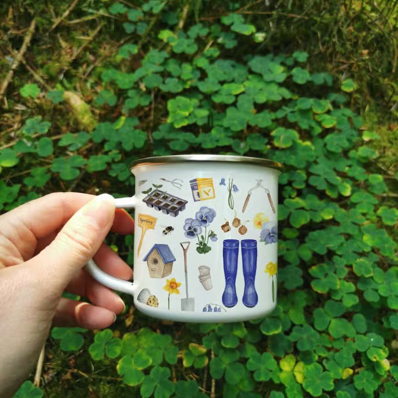 An enamel mug displayed on a patch of clover - the mug has watercolour gardening motifs