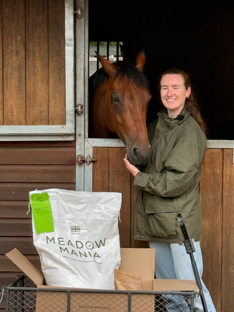 A woman in an olive green jacket stands in a horse stable next to a brown horse, with a large white bag of Meadow Mania horse feed in the foreground.