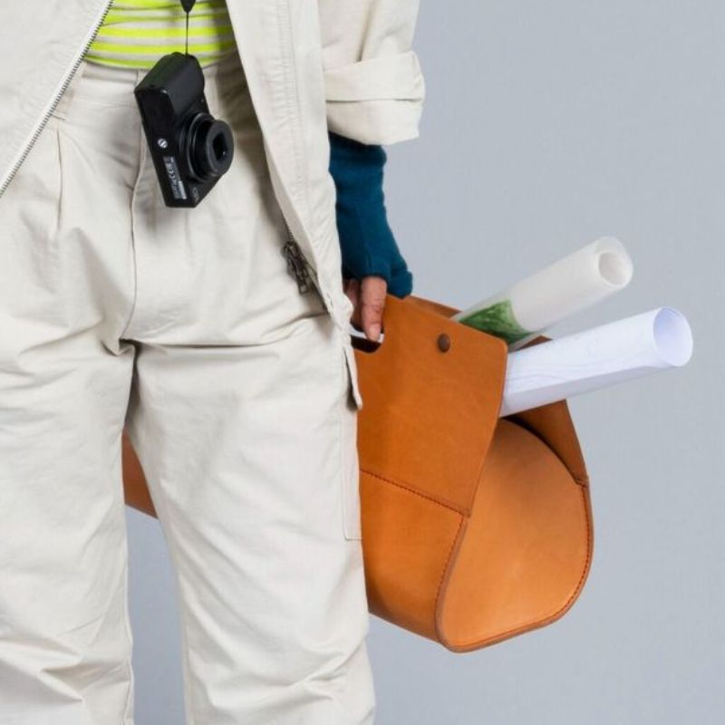 Person holding a brown leather Niwaki tool bag with rolled-up papers against a grey background