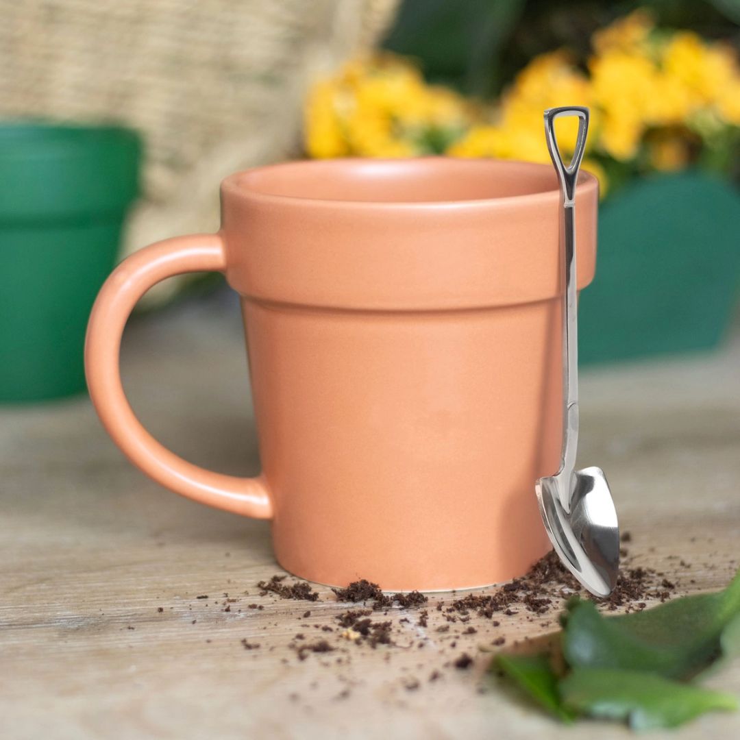 A terracotta-coloured ceramic mug designed to look like a plant pot, accompanied by a mini shovel spoon, displayed on a wooden surface with soil and garden background.