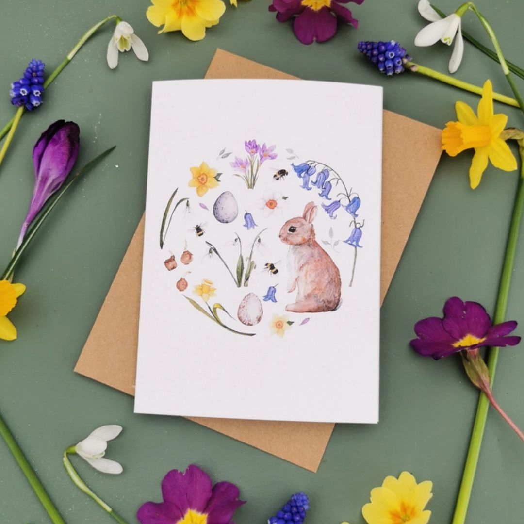 Greeting card with watercolour illustration of a rabbit, some eggs, some insects and wildflowers painted in a circle